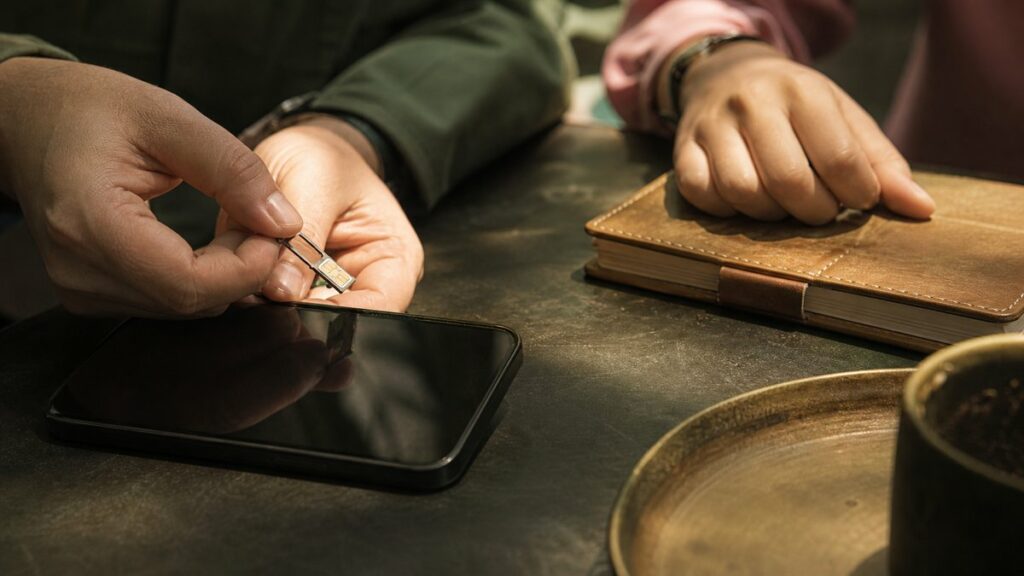 A close hand action scene at a quiet counter where one person gently slides a SIM card into a phone tray while another steadies a small notebook. Warm side light creates calm contrast, with subtle #A98B2D highlights and deep #43544B tones, capturing a thoughtful, grounded sense of confidence.