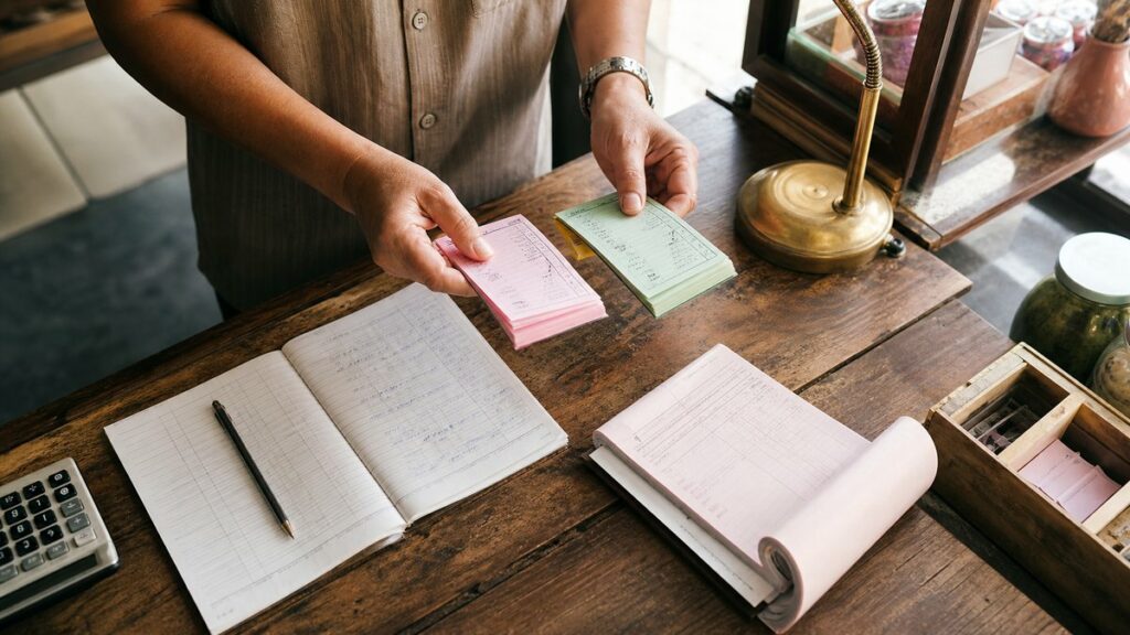 An overhead process photograph of a shop counter where a pair of hands compares printed number cards, a receipt book, and a simple ledger under warm daylight. The composition feels orderly and trustworthy, with natural shadows, a subtle gold accent from a desk lamp, and restrained pink and green tones in the background objects.