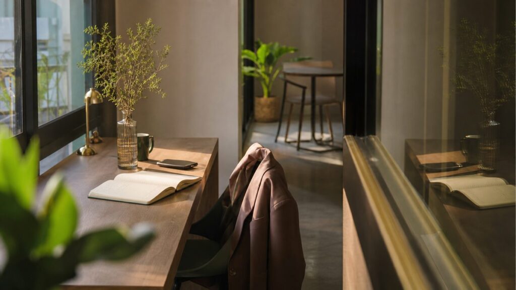 A wide environment view of a quiet co-working corner near a window, where an opened notebook, a phone, and a jacket draped over a chair suggest a fresh start. Late-afternoon light creates soft shadows, while subtle gold and green details add warmth. The atmosphere is focused and optimistic.