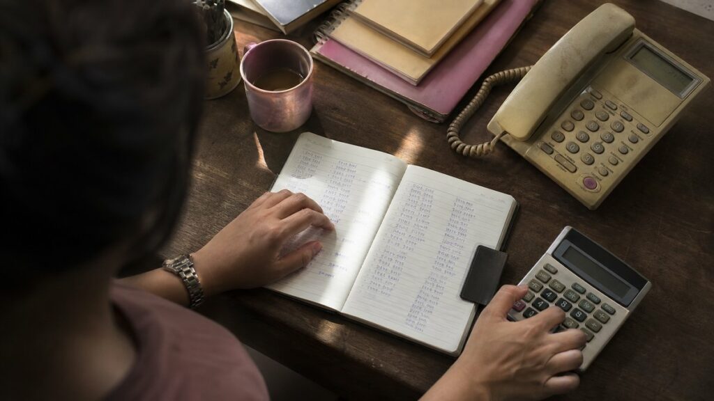 A close hand action scene at a desk shows one hand hovering over a paper notebook filled with neatly written phone numbers, while the other hand taps a calculator beside a worn work phone. Soft window light falls across the scene, with muted rose and olive reflections in the stationery, creating a thoughtful, practical mood.
