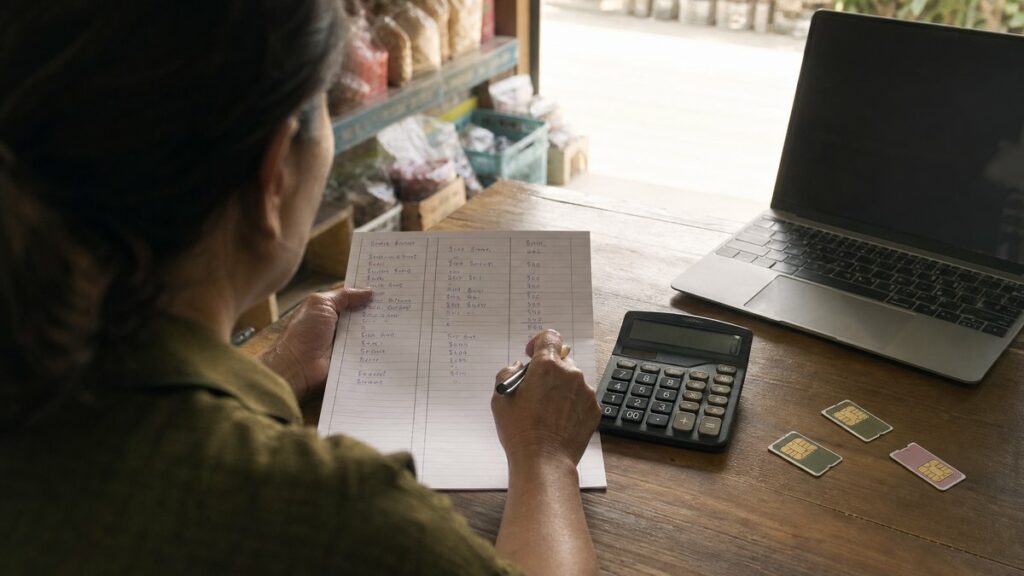 An overhead process shot of a hand comparing a handwritten budget sheet, a calculator, and several SIM cards laid beside a laptop in a modest shop workspace. Natural daylight falls evenly across the table, with restrained rose and olive tones. The mood feels practical, cautious, and organized.