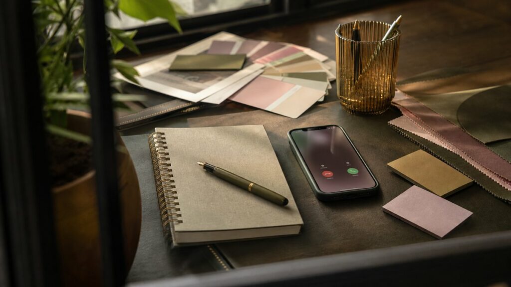 An editorial still life on a designer’s desk: a neatly arranged notebook, color swatches, fountain pen, and business cards beside a softly lit phone displaying an unreadable call screen. Warm window light falls across muted rose, olive, and gold objects, suggesting a creative workflow built on memorable, effortless contact.