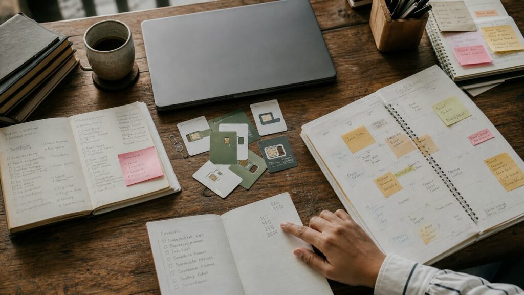 Overhead process scene of a creative planning table with handwritten notes, calendar pages, sticky notes, and several SIM card sleeves spread around a closed laptop. A single hand points between task lists and a small stack of numbers, under diffused daylight, creating a focused, strategic mood.