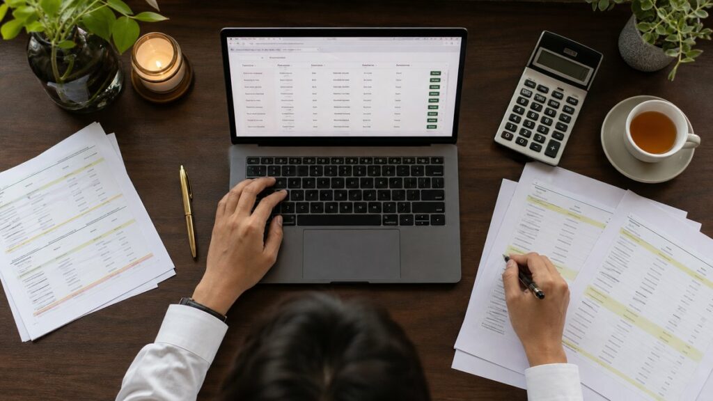 Overhead view of a laptop on a clean workstation displaying a marketplace of phone numbers, surrounded by printed comparison sheets, a calculator, and a cup of tea. Natural window light and orderly composition suggest careful evaluation, while subdued gold and green accents in stationery add a trustworthy, professional tone.