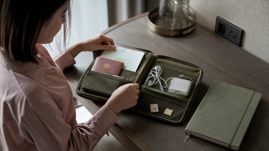 An overhead still life of a neatly packed travel organizer on a hotel desk: passport, folded itinerary, earphones, charger, and two SIM cards beside a sleek notebook. Soft morning light falls across muted pink and olive tones, creating a calm, practical mood that suggests preparation, mobility, and reliable communication.